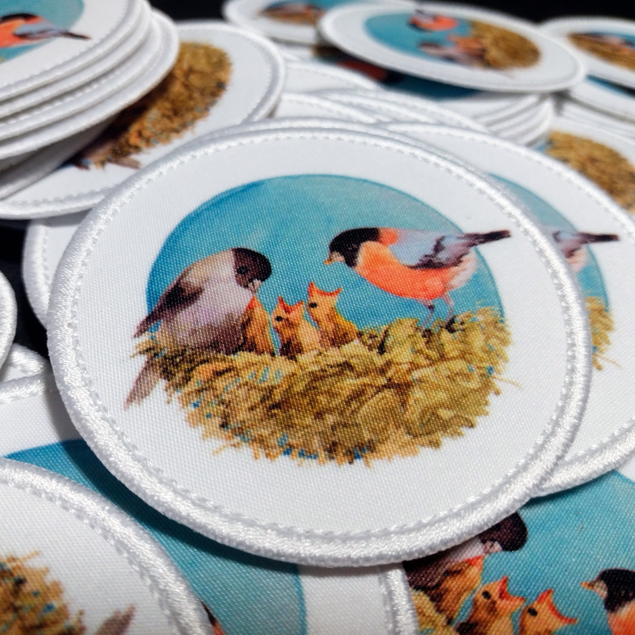 Stos okrągłych naszywek z haftowanym wzorem ptasiej rodziny w gnieździe na błękitnym tle, wykończonych białym, gęstym ściegiem.