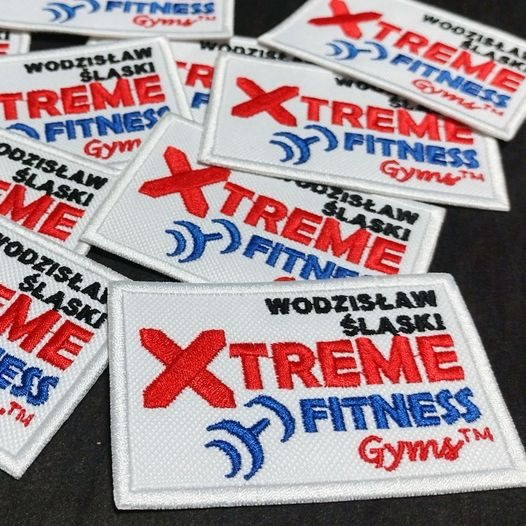 Partia kwadratowych naszywek z haftowanym logo 'Xtreme Fitness Gyms' w kolorach czerwonym i niebieskim na białym tle, ułożonych na czarnym podłożu.