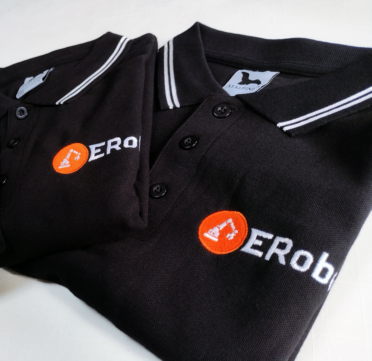 Dwie czarne koszulki polo z haftowanym logo firmy ERoby, logo przedstawia biały napis i pomarańczowe kółko z białym symbolem robota budowlanego, kołnierzyki koszulek wykończone białymi paskami.
