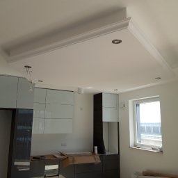 Paul & Zbig Usługi Remontowo - Budowlane Paweł Fijałkowski & Zbigniew Zalewski S.C. - Nowoczesna kuchnia w trakcie wykończenia: białe ściany, modułowe szafki kuchenne z połyskiem, podwieszany sufit z dekoracyjną listwą i oświetleniem punktowym, widoczne kartony i materiały budowlane.