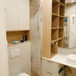 Nowoczesna łazienka z białymi szafkami, drewnianymi półkami i marmurowymi płytkami, wyposażona w wiszącą toaletę i umywalkę nablatową.