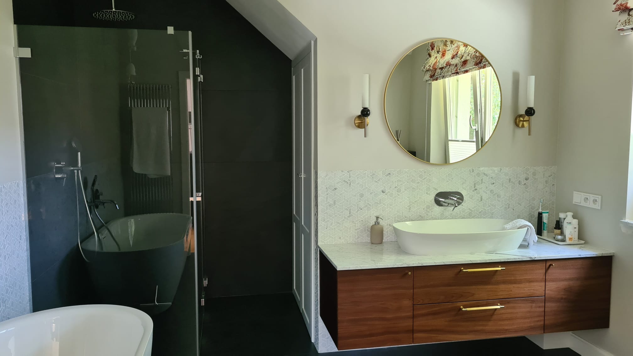 Nowoczesna łazienka z wolnostojącą wanną za szklaną ścianą, okrągłym lustrem z oświetleniem ściennym i umywalką nablatową na drewnianej szafce z marmurowym blatem.