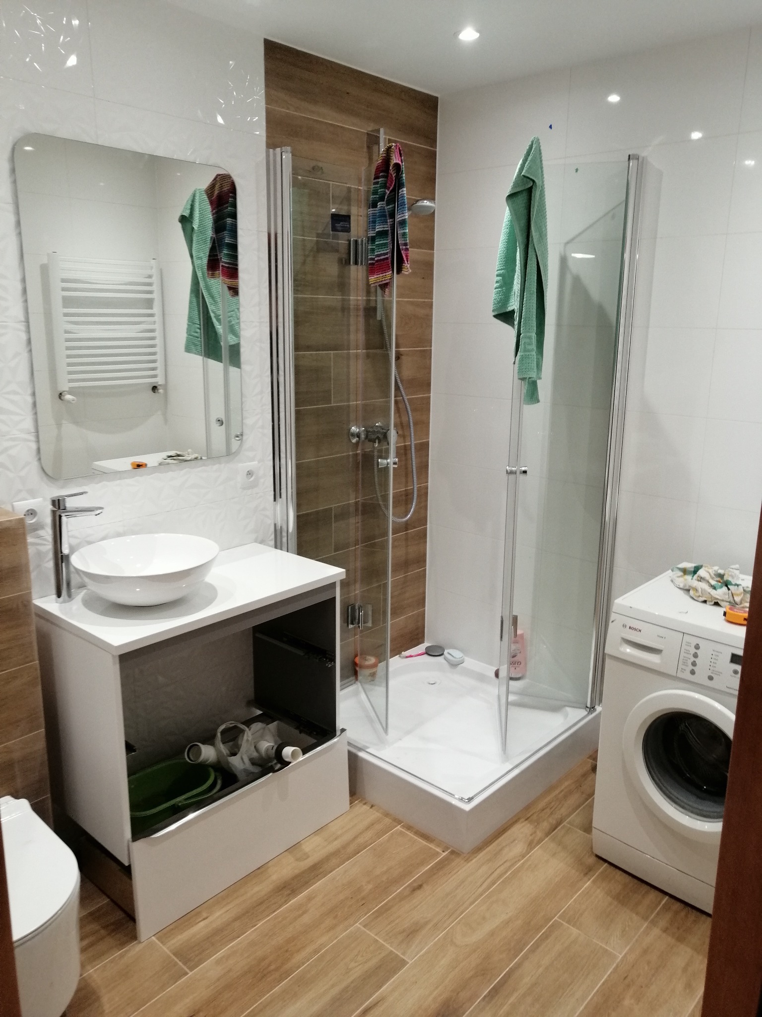 Nowoczesna łazienka z kabiną prysznicową, umywalką nablatową na szafce z otwartą szufladą i pralką, wykończona płytkami w odcieniach bieli i drewna.