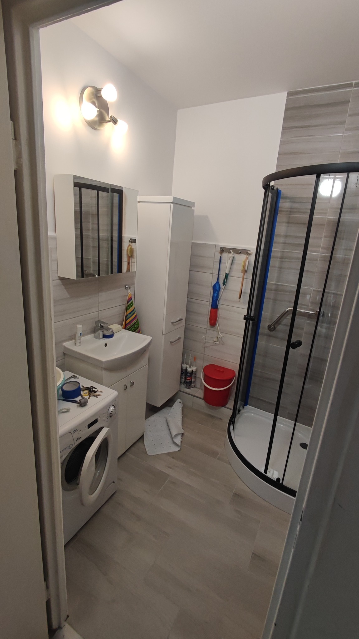 Wnętrze łazienki z kabiną prysznicową, umywalką z szafką, wysoką szafką, pralką i szarą płytką imitującą drewno na ścianach i podłodze.