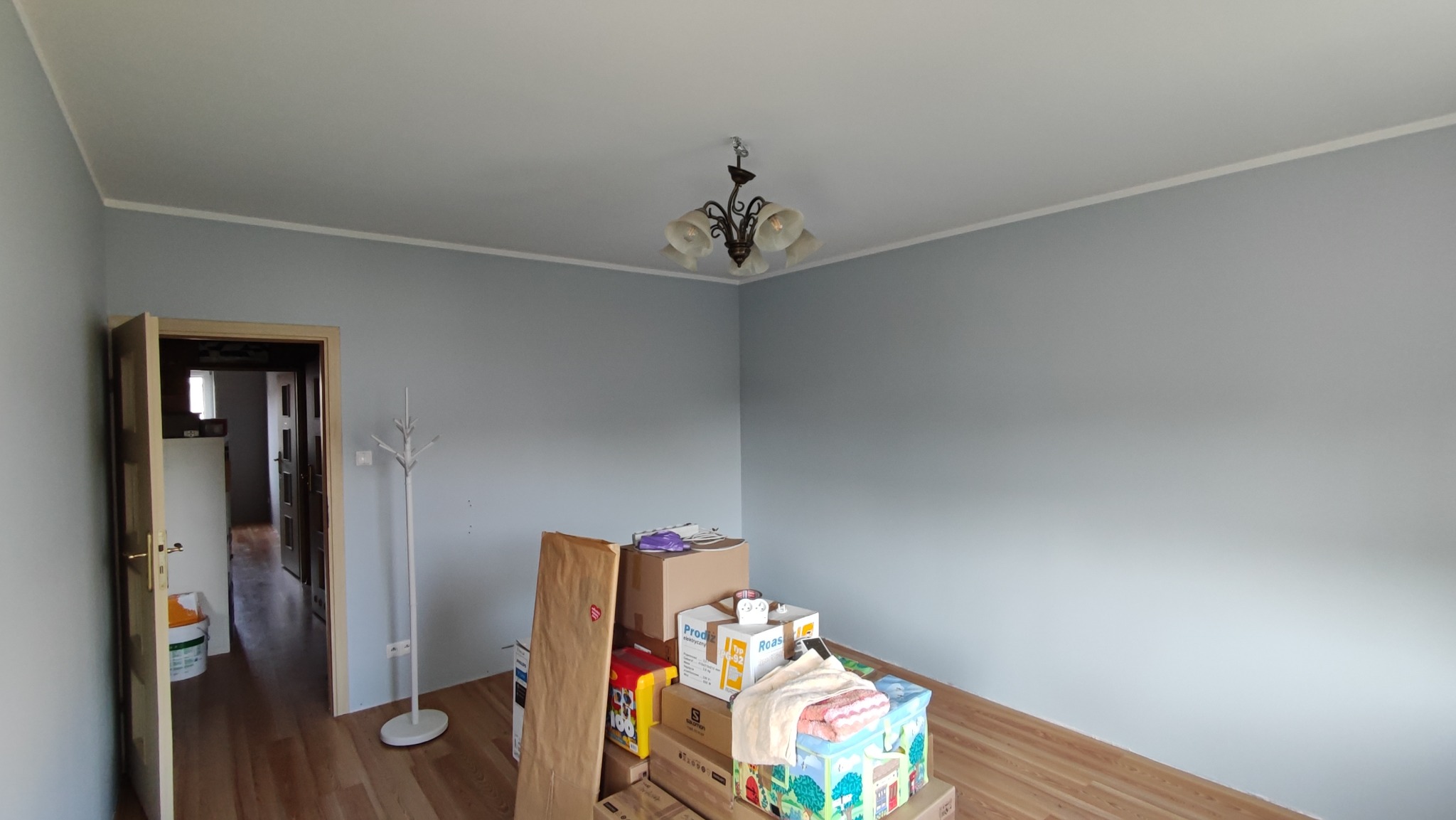 Pomalowane na szaro wnętrze pokoju z podłogą z paneli, z otwartymi drzwiami i stertą kartonów po przeprowadzce, widoczny wieszak na ubrania.