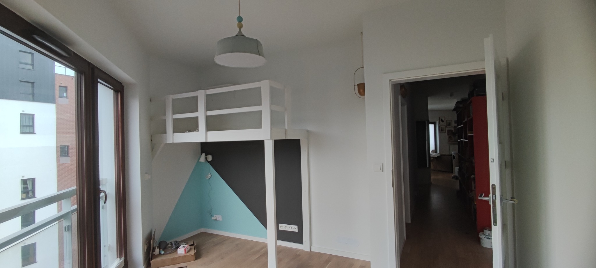 Pokój dziecięcy z łóżkiem na antresoli, ściany pomalowane w geometryczne wzory: trójkąt turkusowy i prostokąt grafitowy, drewniana podłoga, widok na balkon i budynki za oknem.