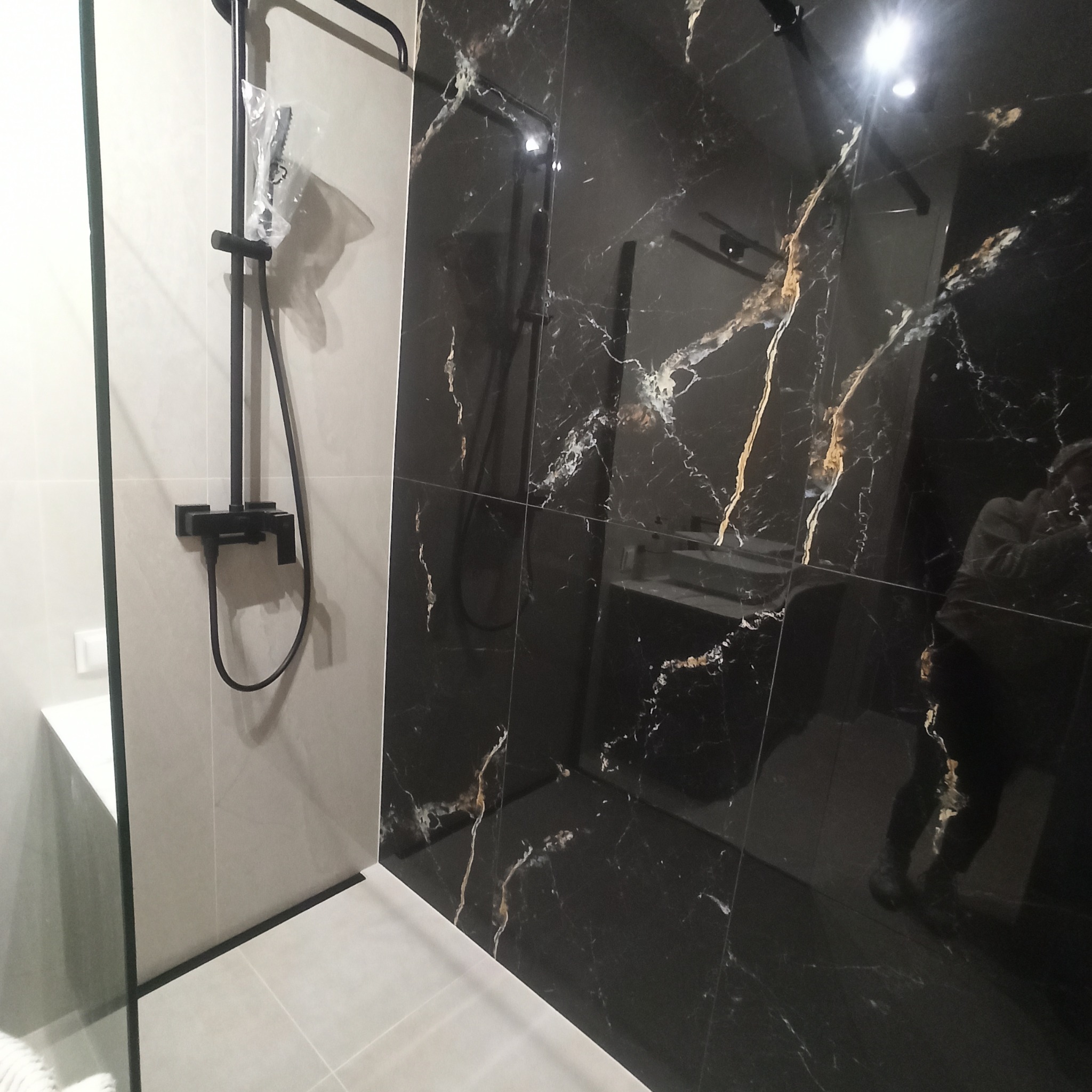 Nowoczesna łazienka z czarnymi płytkami imitującymi marmur z wyraźnymi złotymi żyłami, kontrastującymi z jasnymi, matowymi płytkami i czarną armaturą prysznicową, częściowo jeszcze w folii ochronnej.