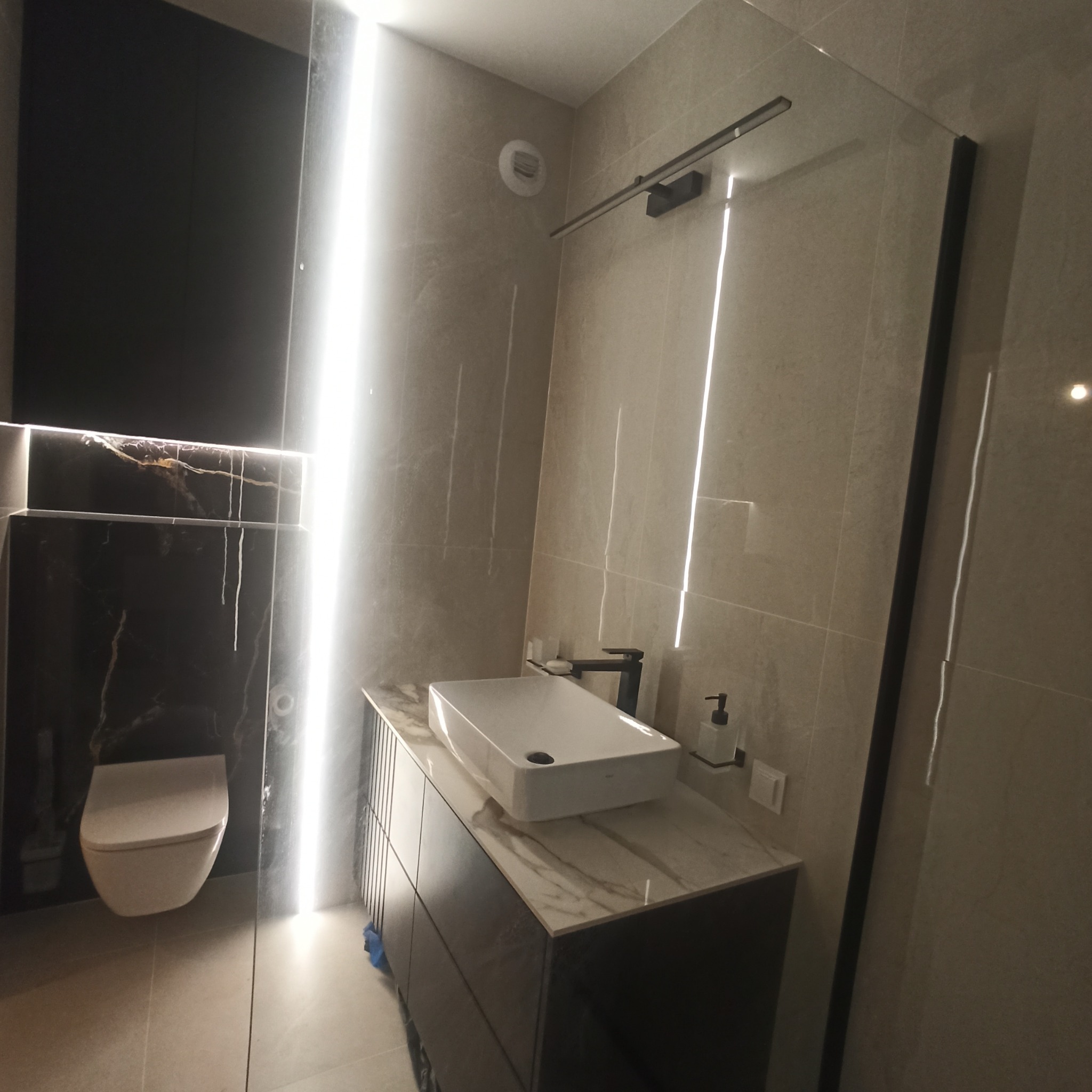 Nowoczesna łazienka z podświetlaną ścianą, umywalką nablatową na szafce z marmurowym blatem, toaleta podwieszana na tle czarnej ściany z marmurowym akcentem, kabina prysznicowa ze szklaną ścianą.