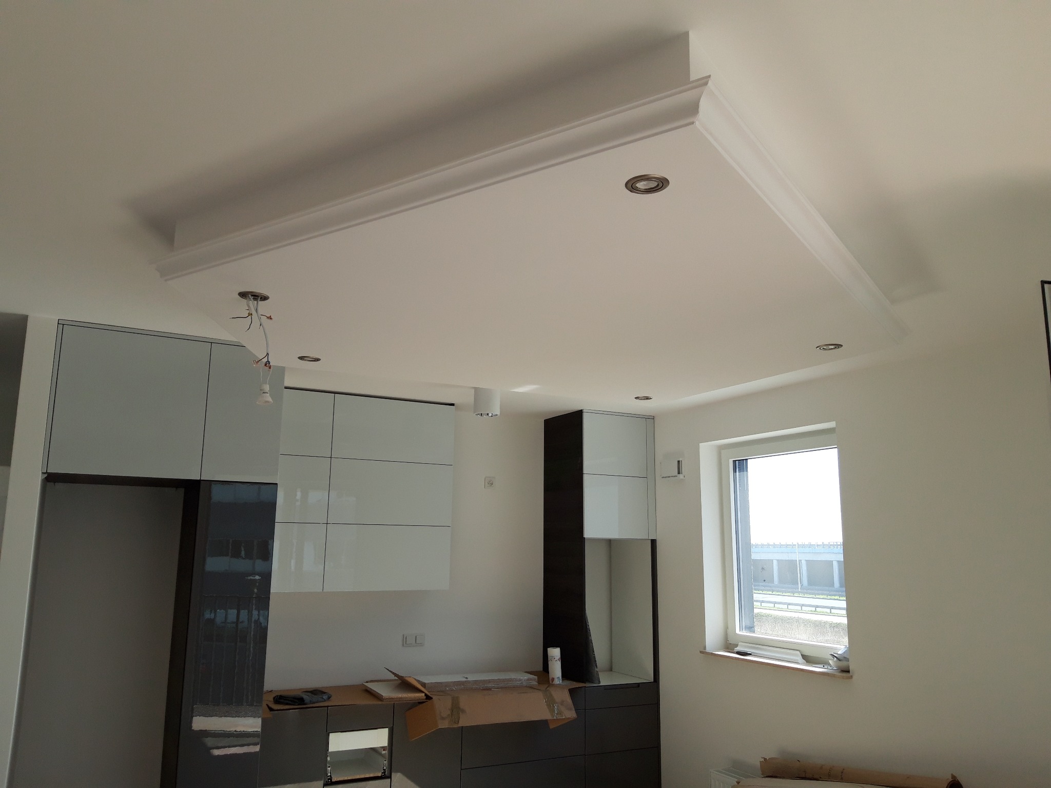 Nowoczesna kuchnia w trakcie wykończenia: białe ściany, modułowe szafki kuchenne z połyskiem, podwieszany sufit z dekoracyjną listwą i oświetleniem punktowym, widoczne kartony i materiały budowlane.