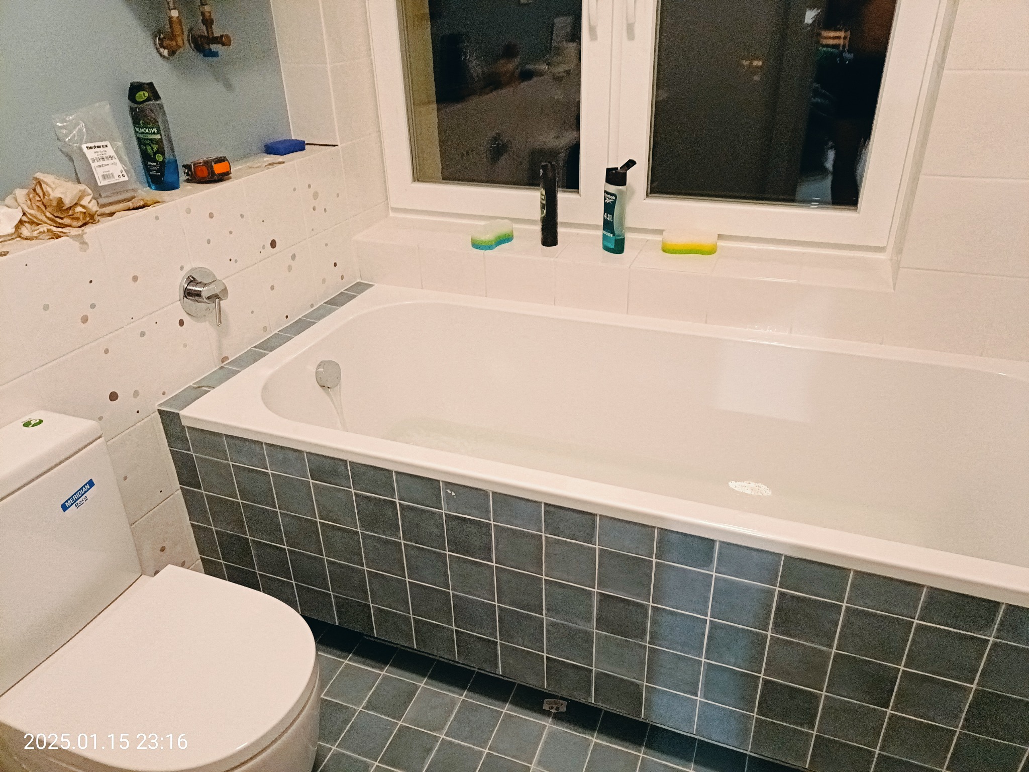 Ukończona łazienka z wanną obłożoną ciemnymi kwadratowymi płytkami, białą toaletą i akcesoriami do kąpieli na parapecie okna.