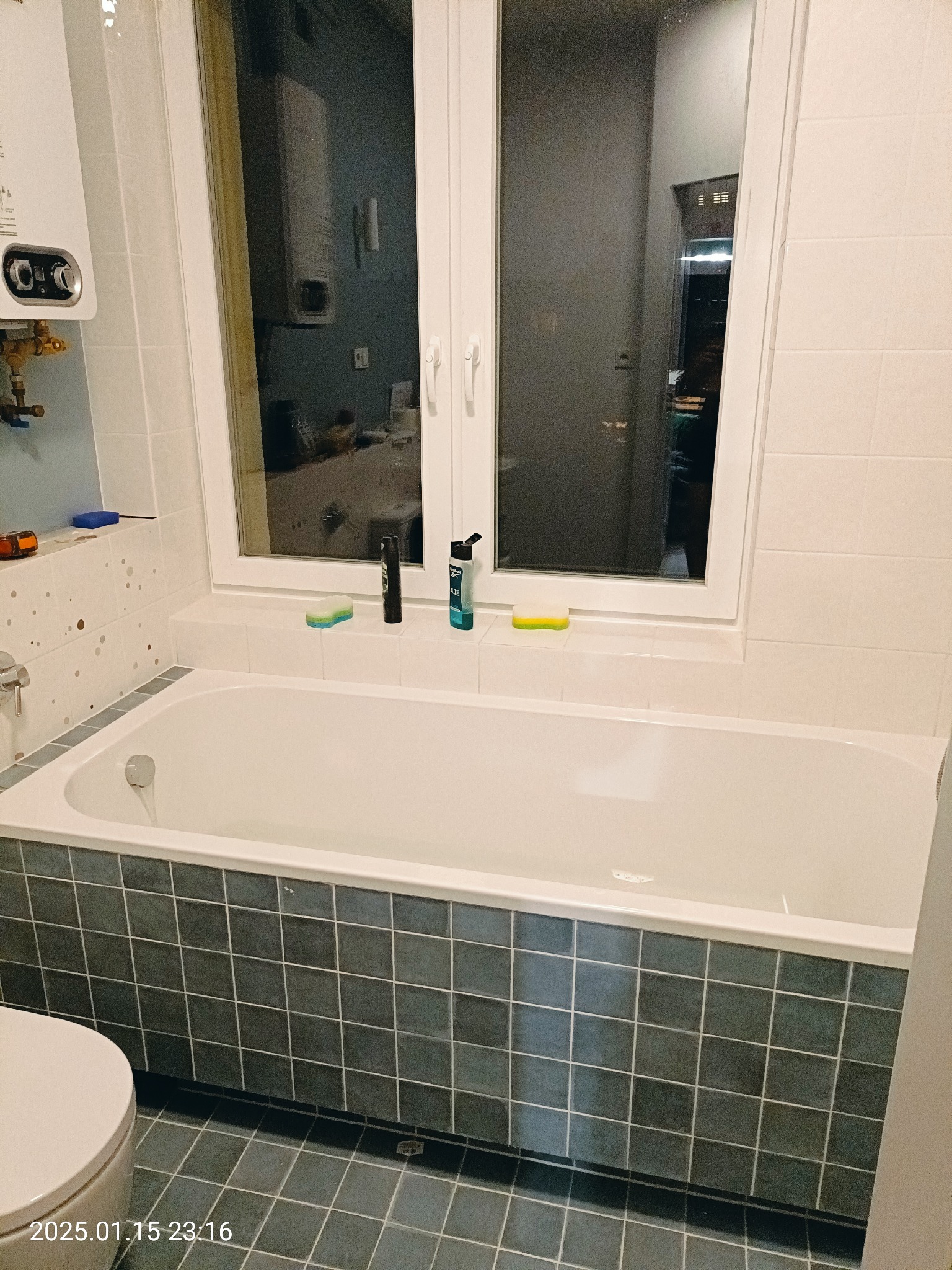 Biała wanna obudowana ciemnymi, kwadratowymi płytkami, z oknem nad wanną i bojlerem gazowym po lewej stronie, widoczne kosmetyki na parapecie, sedes w lewym dolnym rogu.