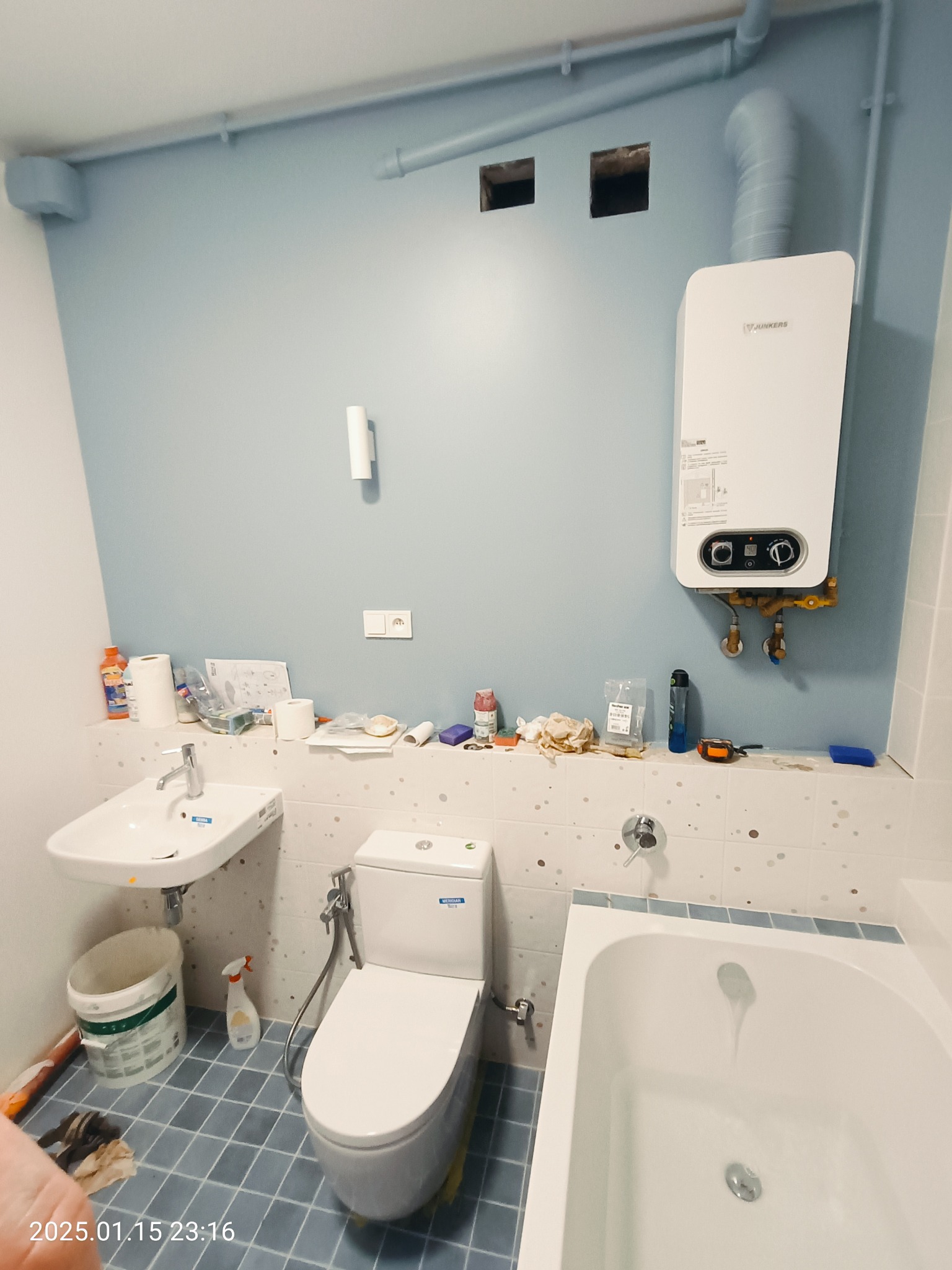 Niedokończona łazienka z niebieskimi ścianami, widoczny bojler Junkers, umywalka, toaleta i wanna. Na półce nad toaletą rozłożone narzędzia i materiały budowlane.