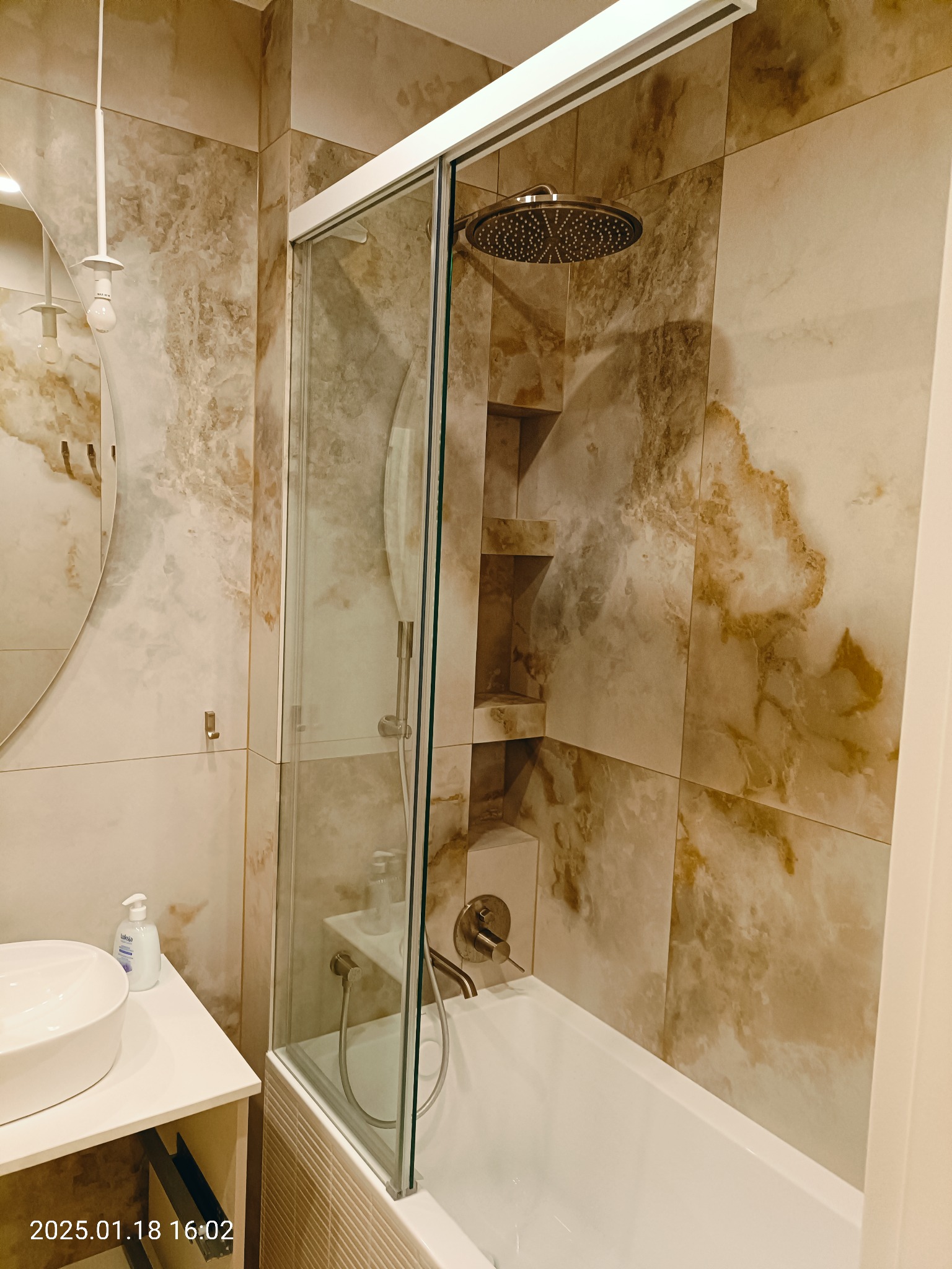 Elegancka łazienka w Warszawie z beżowymi płytkami imitującymi marmur, kabina prysznicowa ze szklanymi drzwiami, okrągłe lustro i umywalka nablatowa.