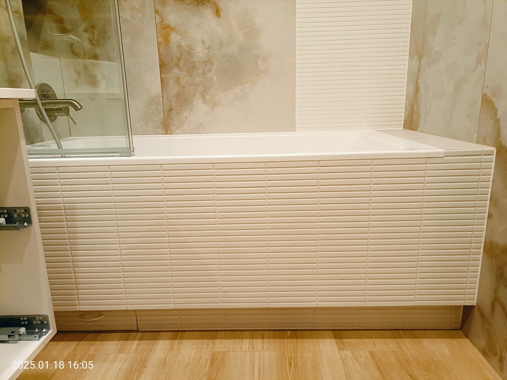 Nowoczesna łazienka z wanną obudowaną drobną, poziomą mozaiką, marmurowe płytki na ścianie i drewnopodobna podłoga, widoczna kabina prysznicowa i fragment szafki z szufladami.