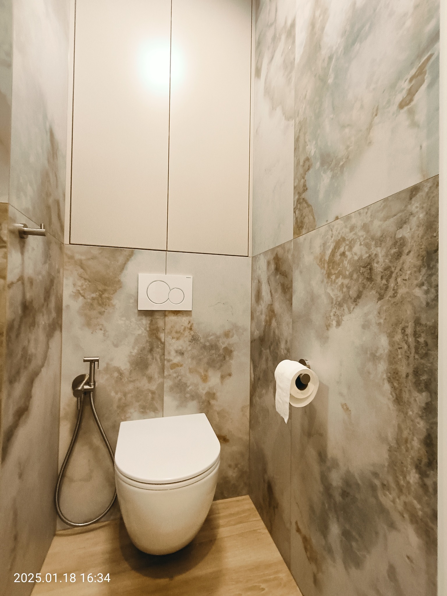 Nowoczesna łazienka z podwieszaną toaletą, bidetką i marmurowymi płytkami ściennymi w odcieniach beżu i szarości, drewnopodobna podłoga.