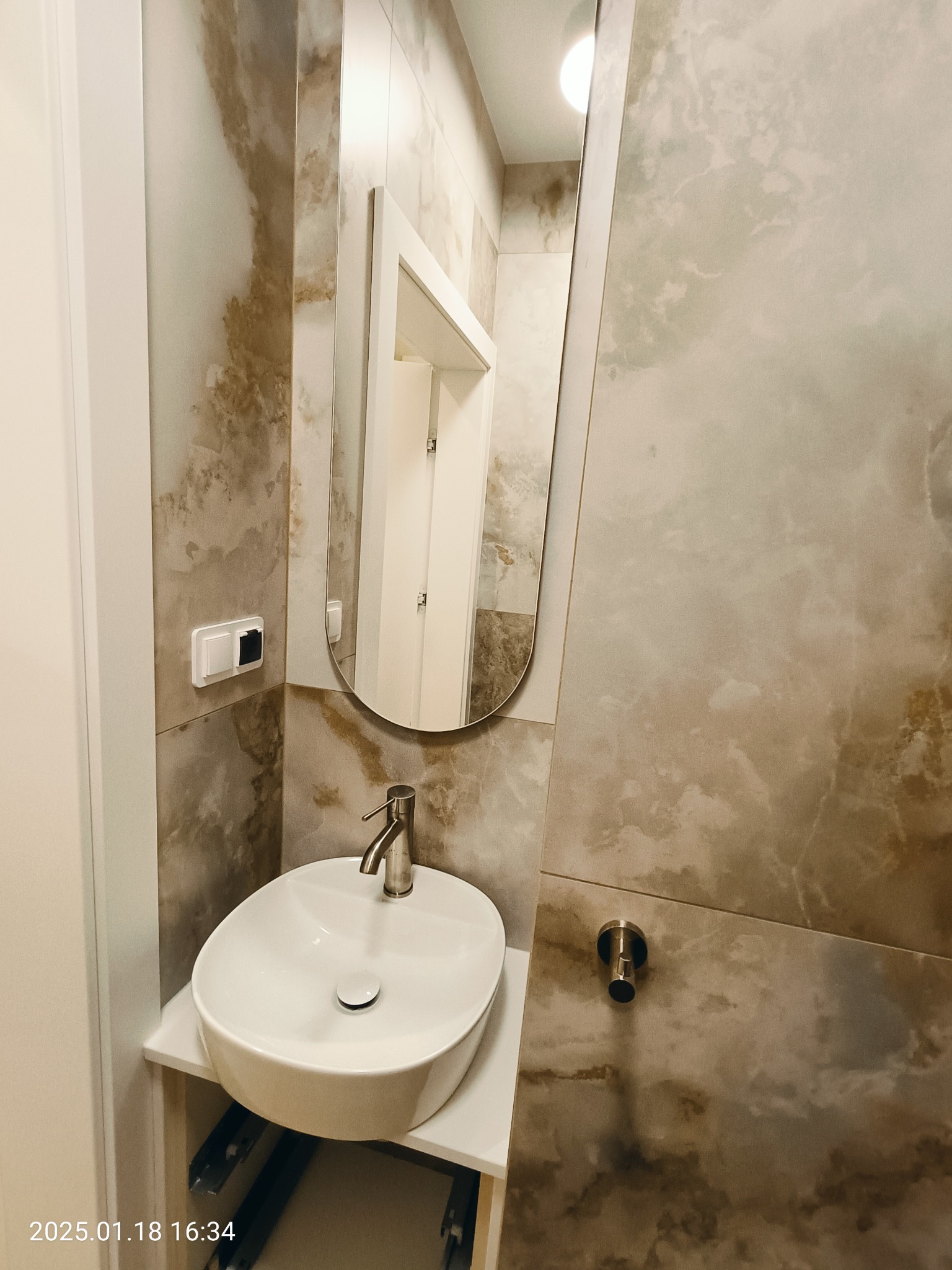 Nowoczesna łazienka z umywalką nablatową, lustrem w owalnej ramie i płytkami imitującymi marmur na ścianach. Widoczny włącznik światła i uchwyt na papier toaletowy.