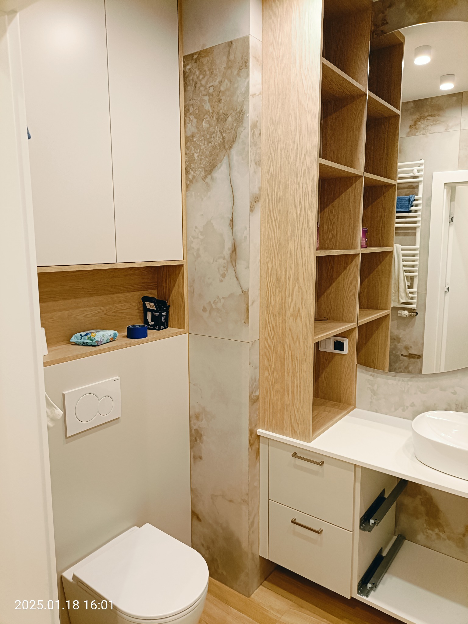 Nowoczesna łazienka z białymi szafkami, drewnianymi półkami i marmurowymi płytkami, wyposażona w wiszącą toaletę i umywalkę nablatową.