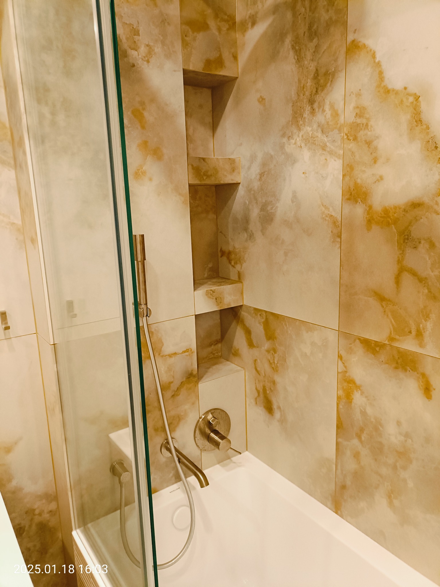 Wykończona łazienka z wanną, szklanym parawanem i armaturą w kolorze szczotkowanego złota, ściany wyłożone płytkami imitującymi marmur, wbudowane półki w ścianie.