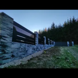 Nowoczesne ogrodzenie z betonowych bloczków o wzorze imitującym kamień i czarnych, poziomych paneli, otoczone jasnymi kamykami, na tle iglastego lasu i błękitnego nieba.