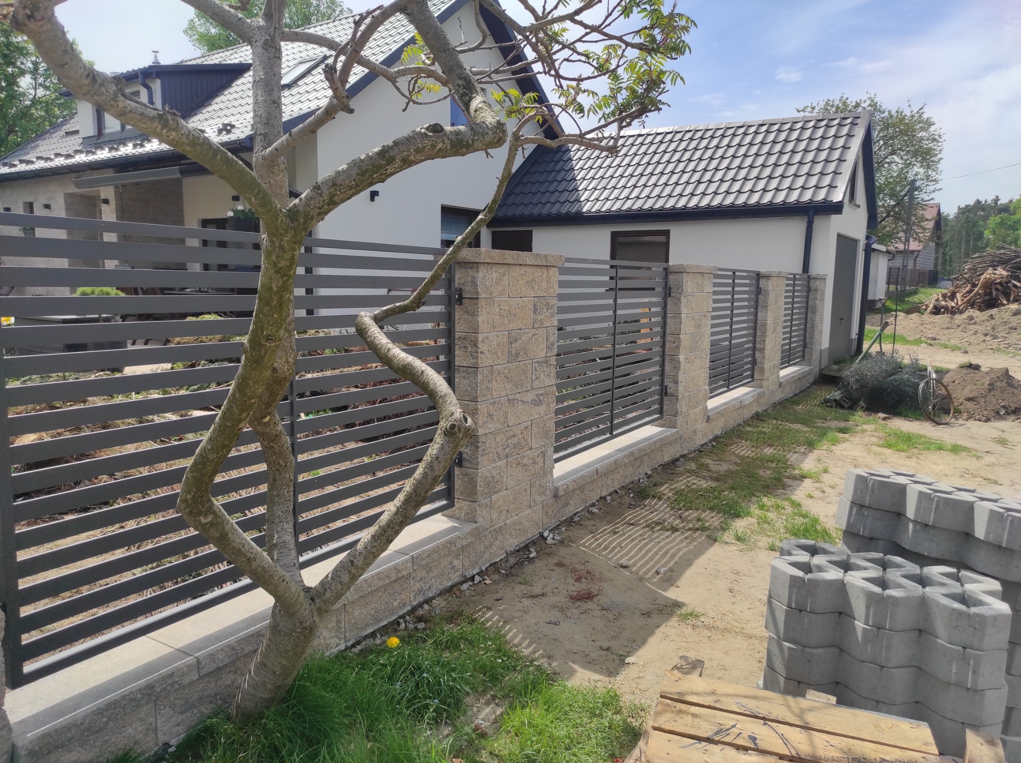 Nowoczesne, poziome przęsła w kolorze grafitowym, tworzące ogrodzenie posesji w Bochni, z kamiennymi słupkami i podmurówką, częściowo zasłonięte przez gałęzie drzewa.
