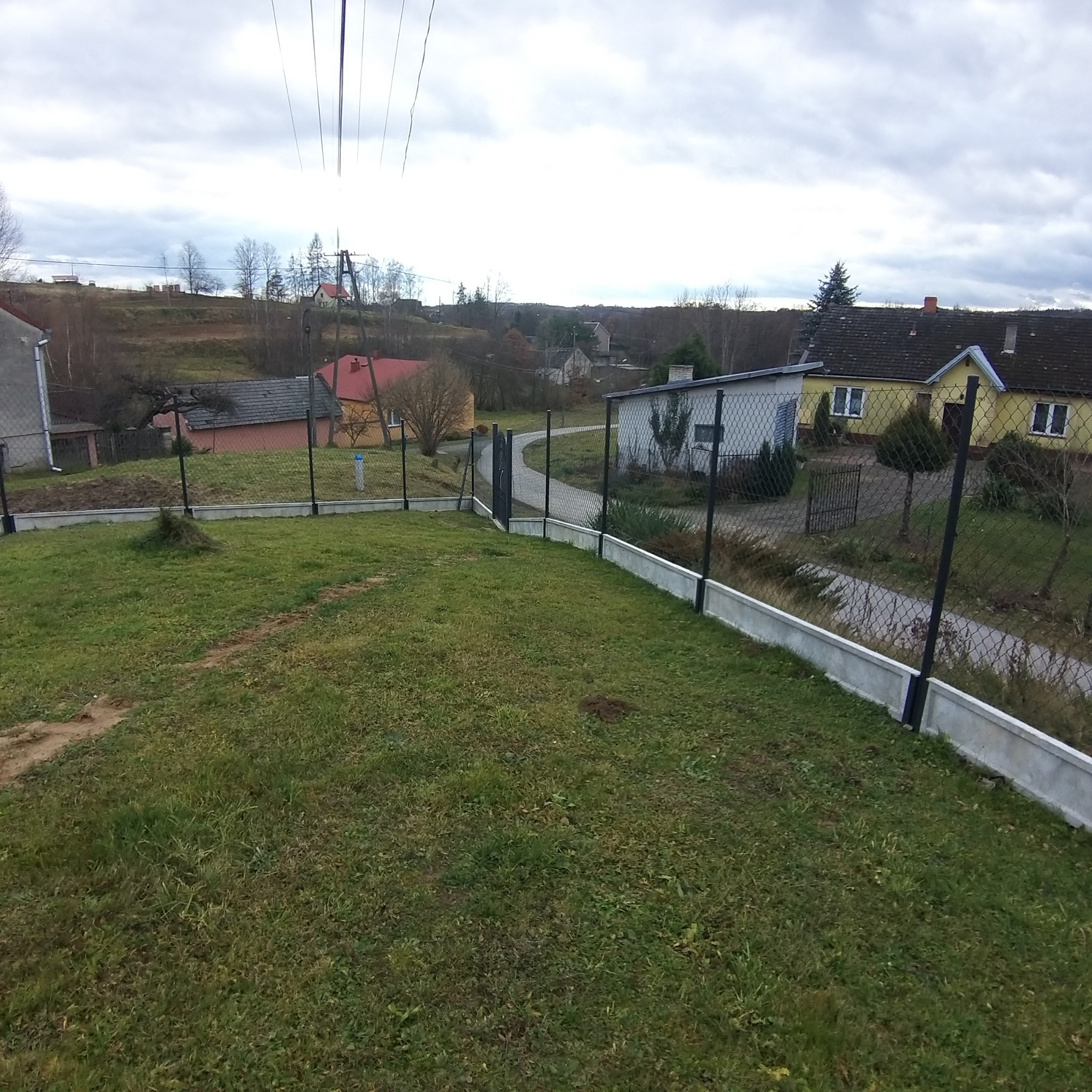 Nowo postawione ogrodzenie z siatki z podmurówką betonową na nierównym terenie, widok na domy w oddali w pochmurny dzień.