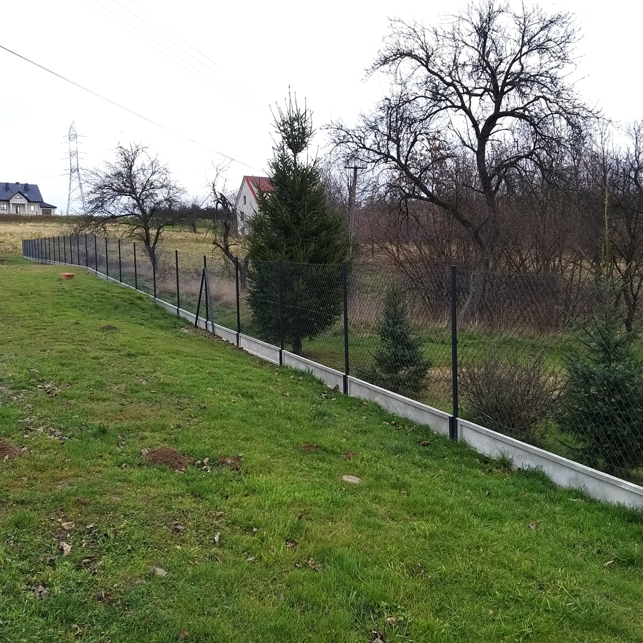 Nowoczesne ogrodzenie z siatki z podmurówką betonową na nierównym terenie, w tle dom jednorodzinny i linia wysokiego napięcia.