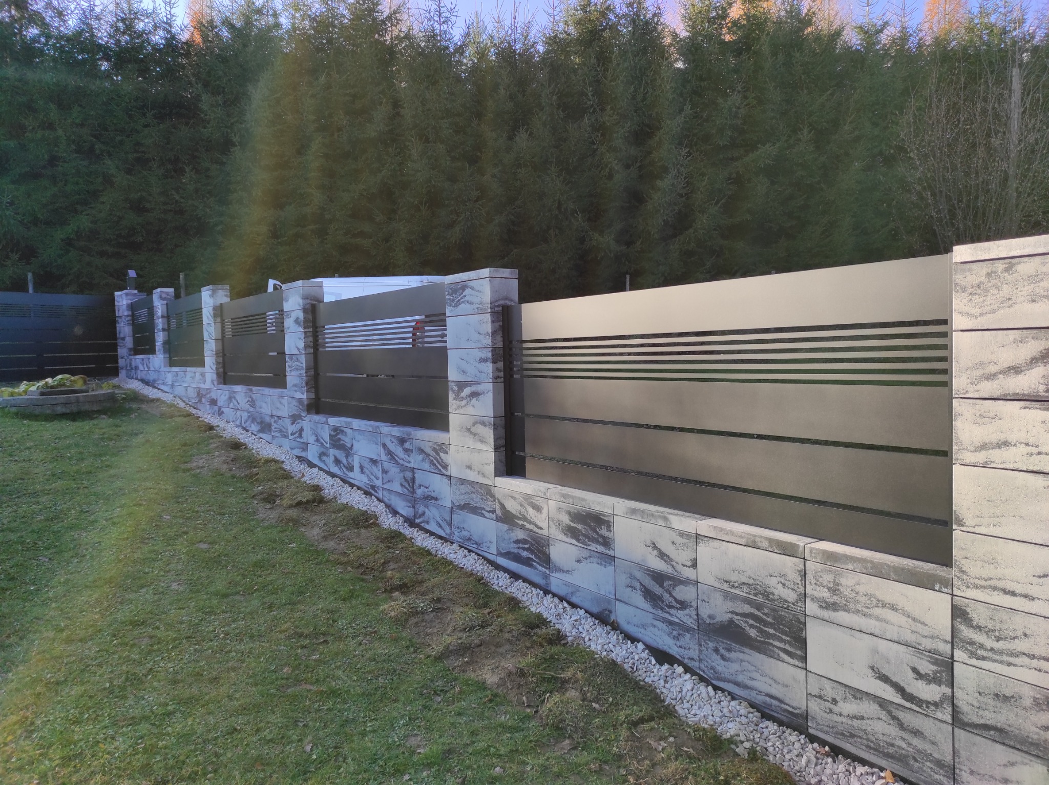 Nowoczesne ogrodzenie z poziomych, szarych paneli metalowych, osadzone na murku z szaro-białych bloczków betonowych, z widocznym pasem białego żwiru u podstawy i zielonym trawnikiem w tle, z rzędem...