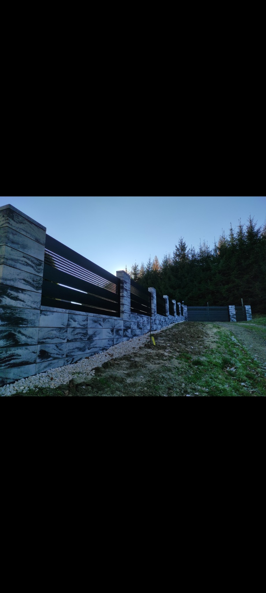 Nowoczesne ogrodzenie z betonowych bloczków o wzorze imitującym kamień i czarnych, poziomych paneli, otoczone jasnymi kamykami, na tle iglastego lasu i błękitnego nieba.