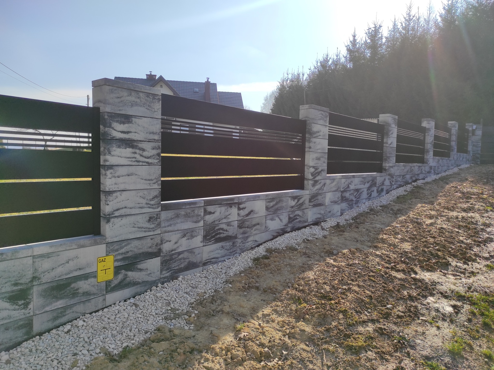 Nowoczesne ogrodzenie z poziomych, czarnych paneli zamocowanych między szarymi, betonowymi słupkami i podmurówką, z widocznym znakiem 'GAZ' na jednym z elementów, otoczone żwirem i ziemią.