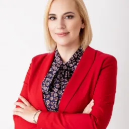 Portret kobiety w czerwonej marynarce i wzorzystej bluzce, z rękami skrzyżowanymi, na jasnym tle.