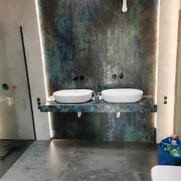 Nowoczesna łazienka z dwoma białymi umywalkami na blacie wykończonym mozaiką, ściana w odcieniach szarości i błękitu, widoczna surowa żarówka i niebieska torba z akcesoriami.