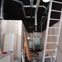 Vent klim - Wnętrze w trakcie remontu z widocznymi instalacjami wentylacyjnymi pod sufitem, aluminiową drabiną opartą o ścianę, przygotowanymi do montażu drzwiami i narzędziami.