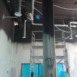 Vent klim - Wnętrze w trakcie prac wykończeniowych: widoczne rury wentylacyjne pod sufitem, ściany w trakcie szpachlowania, zabezpieczone elementy folią ochronną i rozstawiona drabina. Przewody elektryczne...