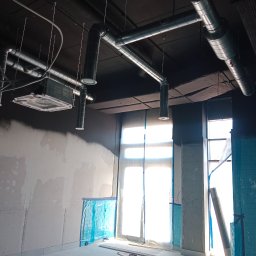 Vent klim - Wnętrze w trakcie remontu z widoczną instalacją wentylacyjną pod sufitem, zabezpieczonymi oknami folią i surowymi ścianami z naniesionymi notatkami.