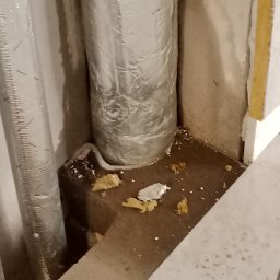 Vent klim - Dwa pionowe przewody wentylacyjne w srebrnej izolacji, usytuowane w rogu pomieszczenia, widoczne ślady pianki montażowej i resztek materiałów budowlanych na podłodze i ścianach.