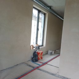 Vent klim - Surowy stan wnętrza z szarymi ścianami, widoczne rury instalacyjne w kolorze czerwonym i niebieskim na podłodze, oraz metalowa rura wentylacyjna pod sufitem, okno z widokiem na zewnątrz, kartonowe...