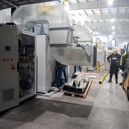 Vent klim - Wnętrze hali produkcyjnej z systemem wentylacyjnym w trakcie instalacji, widoczne kanały wentylacyjne, szafa sterownicza i paleta z elementami montażowymi, dwóch mężczyzn w tle.