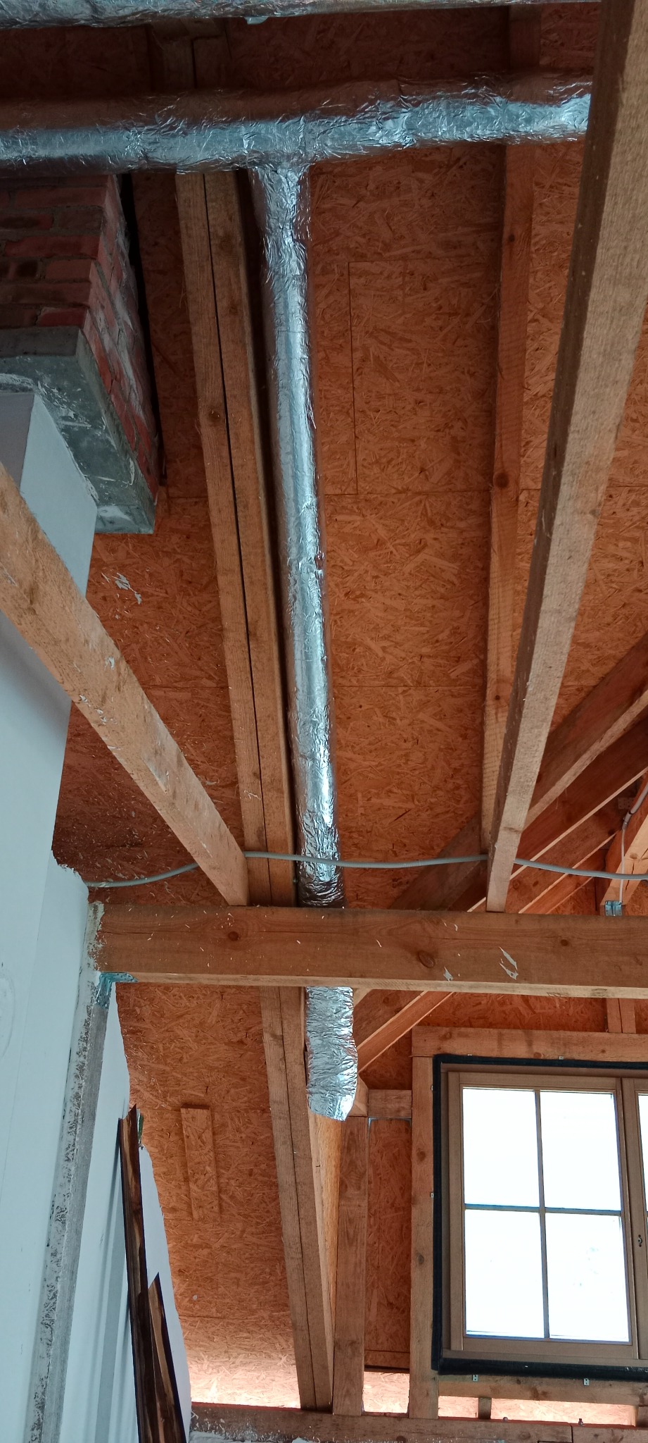 Widok poddasza w trakcie prac: surowa konstrukcja drewniana, płyta OSB na ścianach, rura wentylacyjna w srebrnej izolacji, okno z czarną uszczelką.