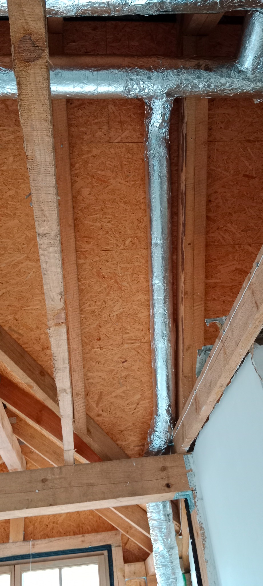 Montaż izolowanego przewodu wentylacyjnego w przestrzeni poddasza z widoczną drewnianą konstrukcją dachu i płytą OSB.