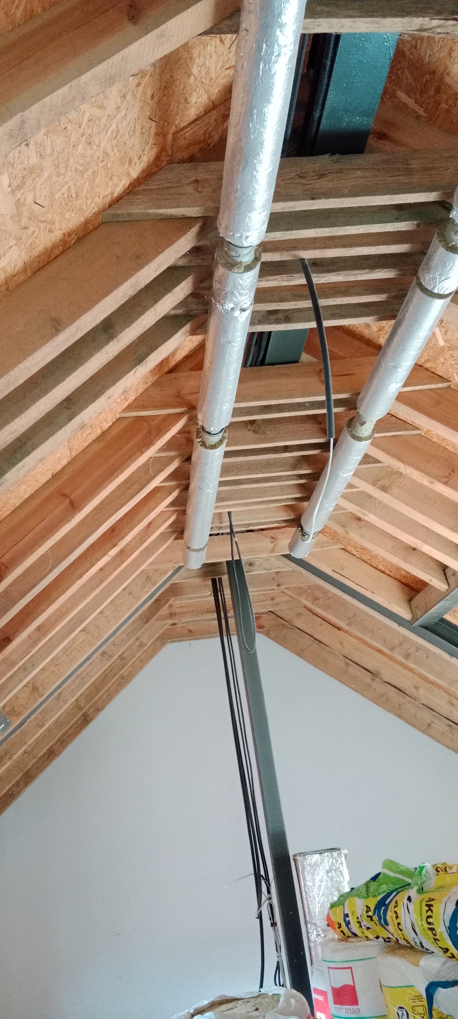 Izolowane rury wentylacyjne pod skośnym dachem z drewnianą konstrukcją więźby, widok z dołu.