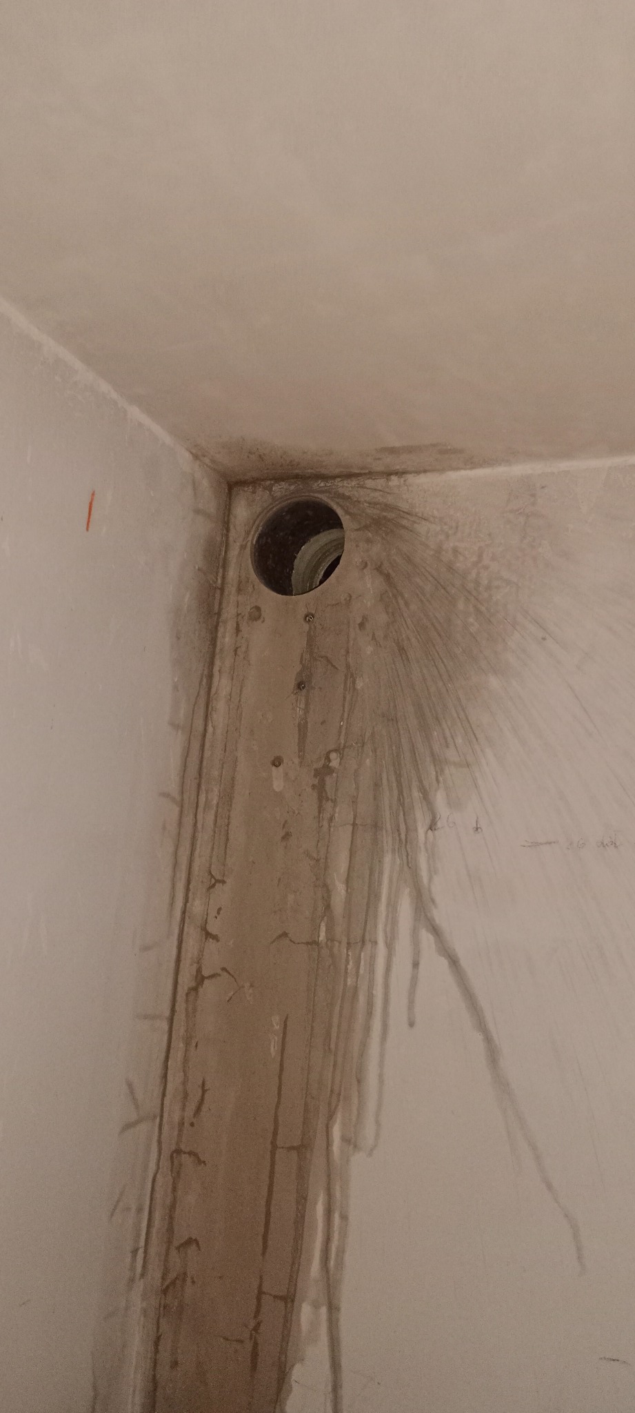 Niedokończony otwór wentylacyjny w rogu pomieszczenia, z widocznymi zaciekami i śladami po wierceniu na surowej ścianie.