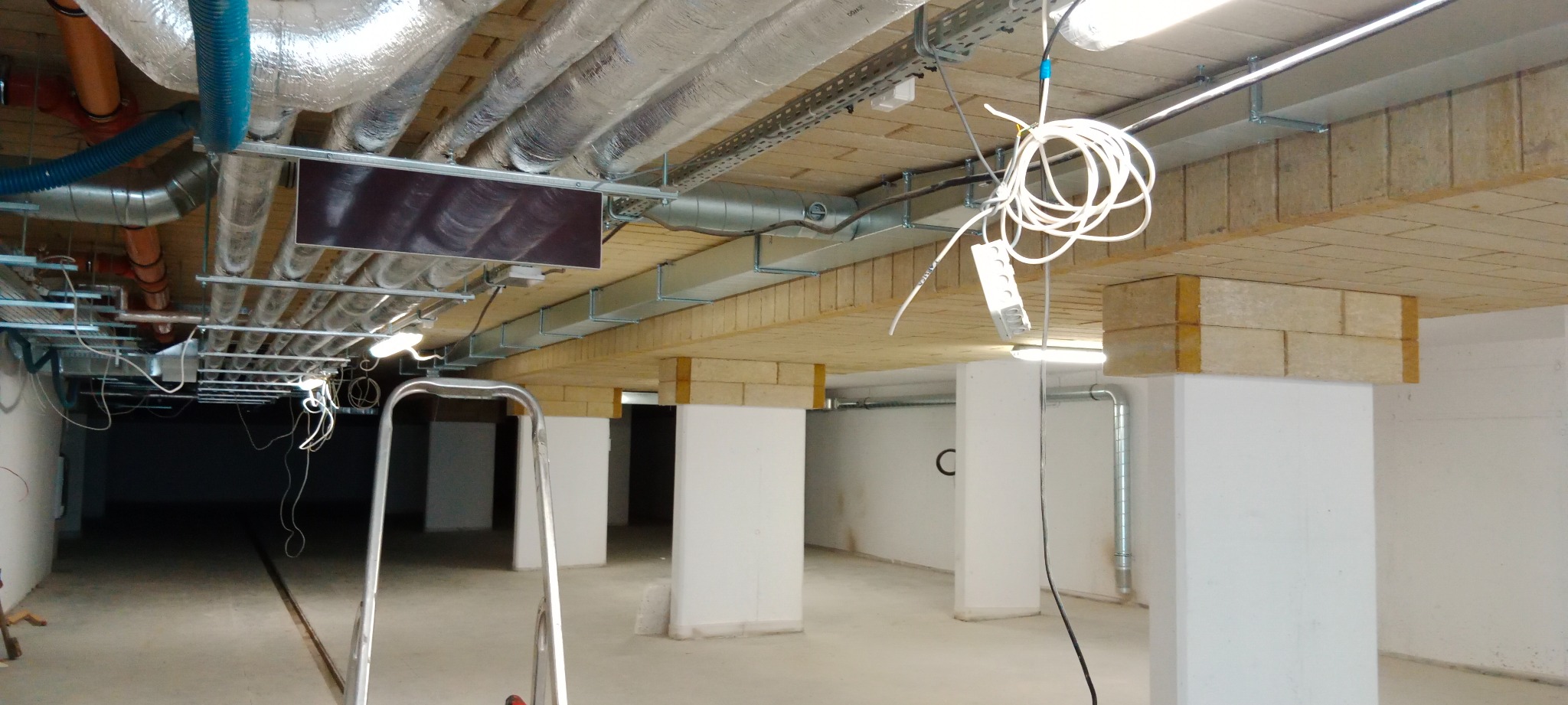 Instalacja wentylacyjna z izolacją termiczną oraz rurami w nowym budynku komercyjnym, widoczne kable i drabina.