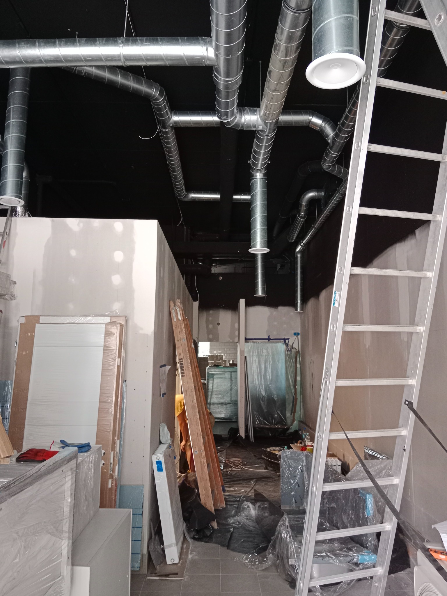 Wnętrze w trakcie remontu z widocznymi instalacjami wentylacyjnymi pod sufitem, aluminiową drabiną opartą o ścianę, przygotowanymi do montażu drzwiami i narzędziami.