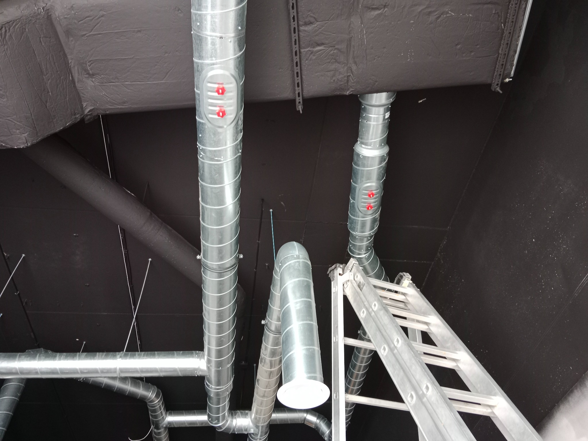 Montaż spiralnych kanałów wentylacyjnych z rewizjami i anemostatem na czarnym suficie, widoczna drabina aluminiowa.