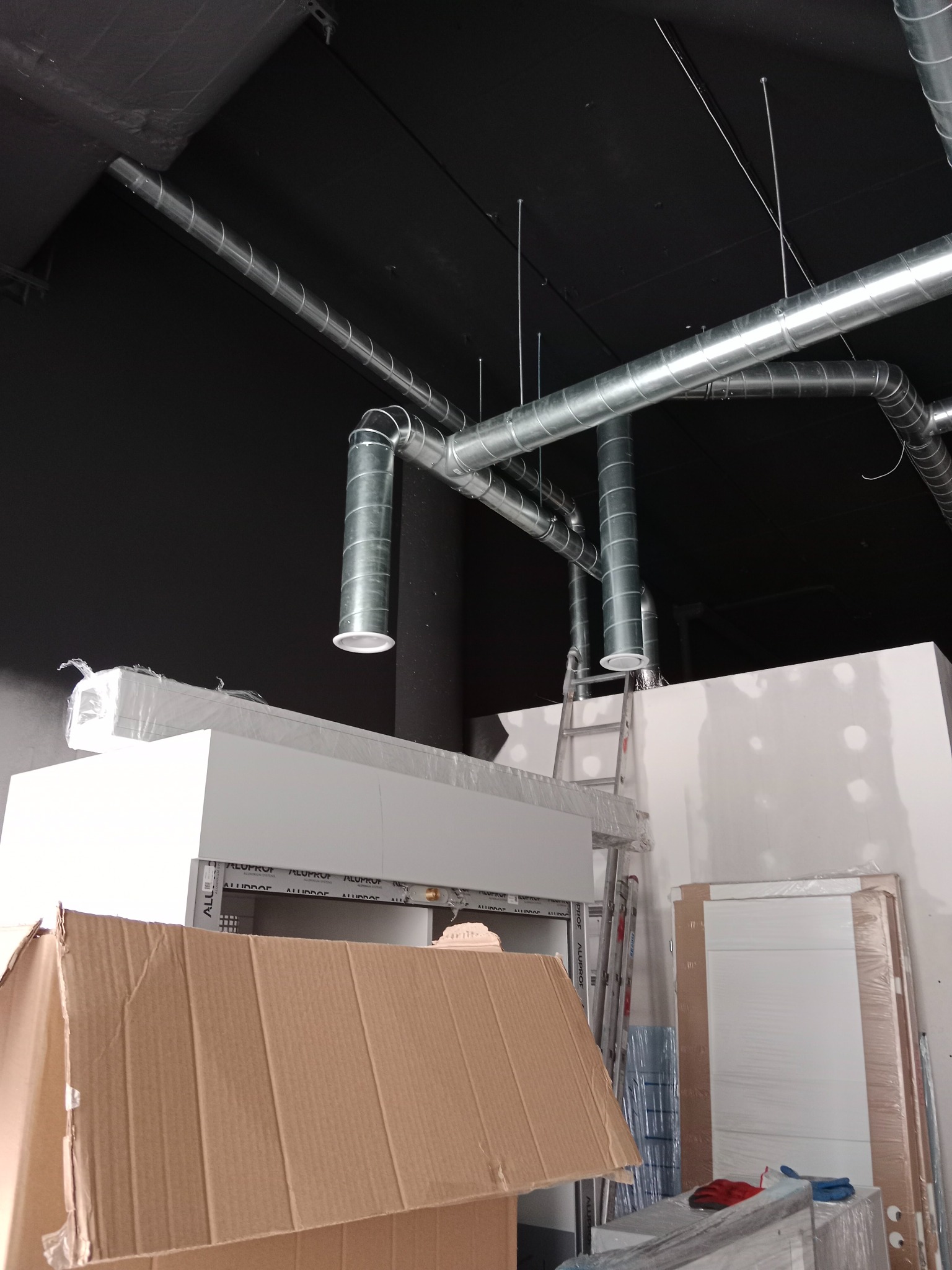 Montaż spiralnych przewodów wentylacyjnych pod czarnym sufitem w budynku komercyjnym, widoczne drabiny, kartony i elementy wykończeniowe.