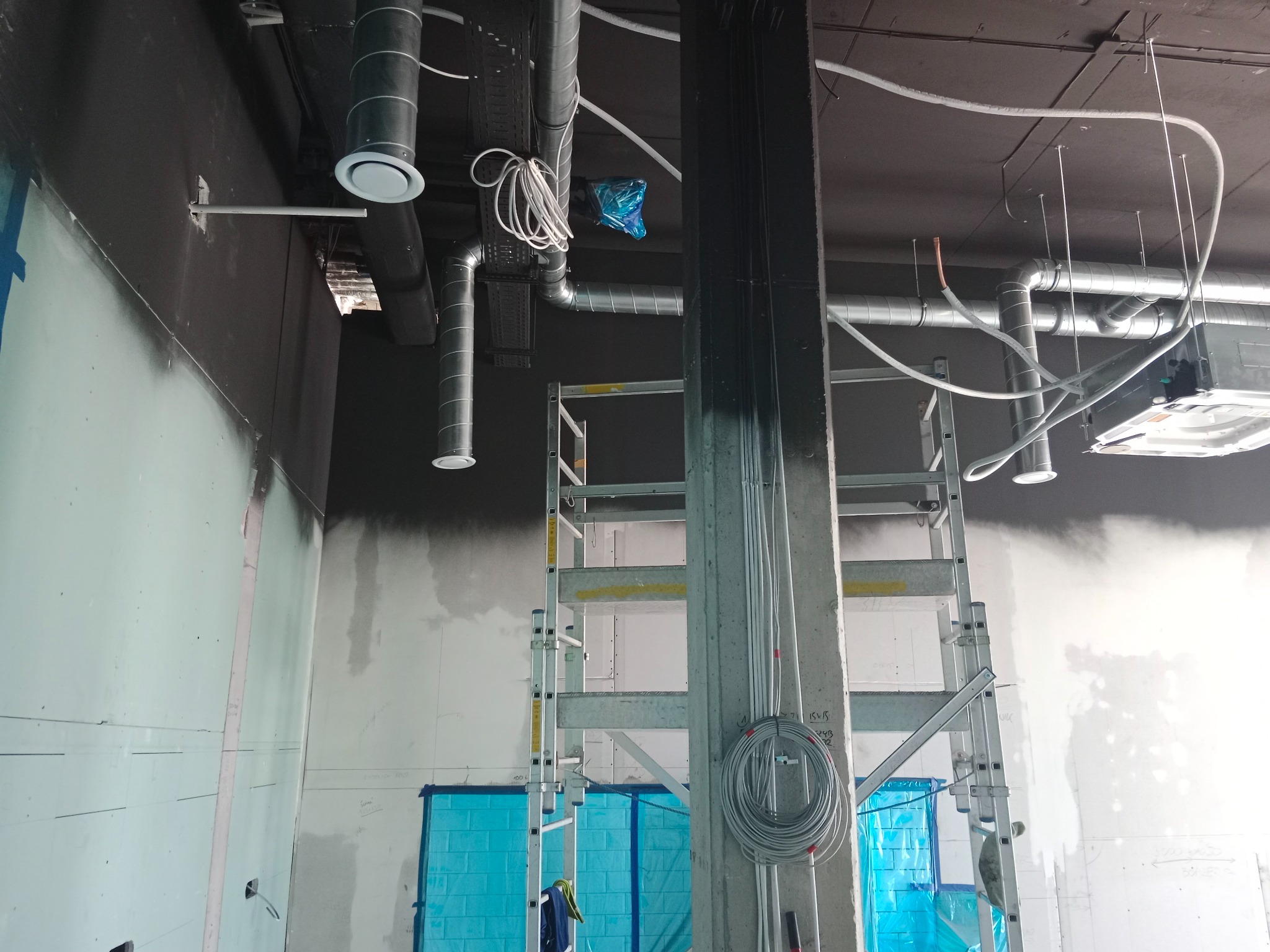 Wnętrze w trakcie prac wykończeniowych: widoczne rury wentylacyjne pod sufitem, ściany w trakcie szpachlowania, zabezpieczone elementy folią ochronną i rozstawiona drabina. Przewody elektryczne...