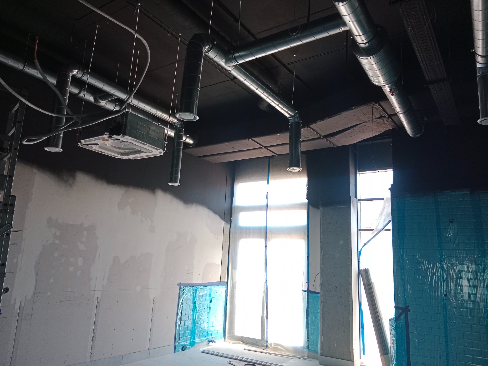 Wnętrze w trakcie remontu z widoczną instalacją wentylacyjną pod sufitem, zabezpieczonymi oknami folią i surowymi ścianami z naniesionymi notatkami.