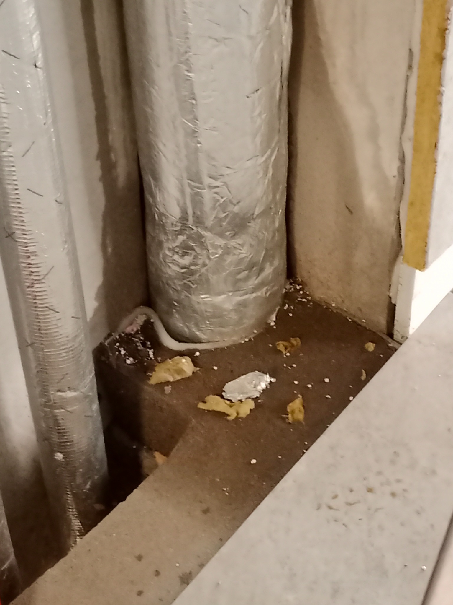Dwa pionowe przewody wentylacyjne w srebrnej izolacji, usytuowane w rogu pomieszczenia, widoczne ślady pianki montażowej i resztek materiałów budowlanych na podłodze i ścianach.