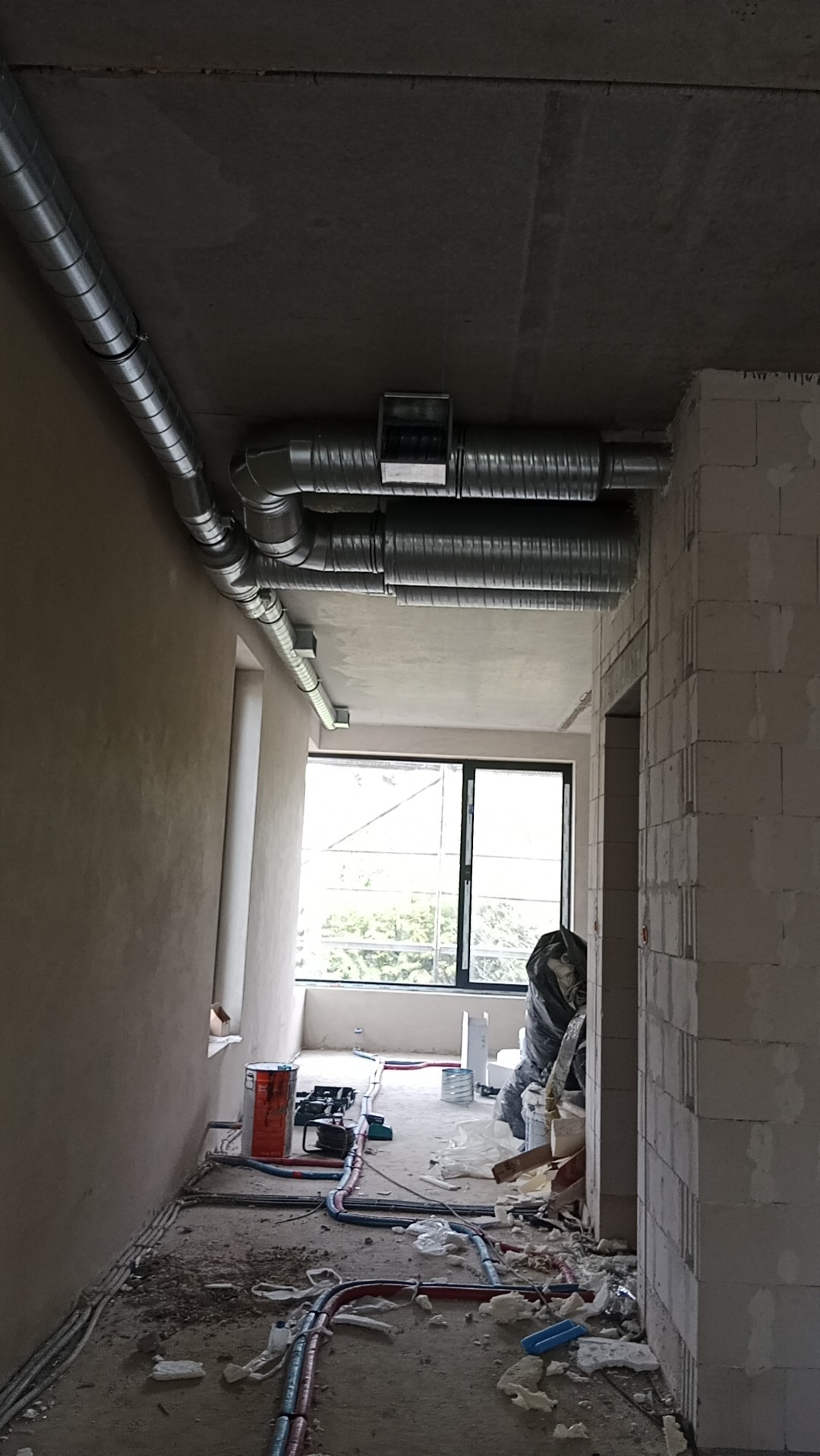 Wnętrze w trakcie montażu instalacji wentylacyjnej z widocznymi przewodami wentylacyjnymi pod sufitem, surowymi ścianami i narzędziami na podłodze.