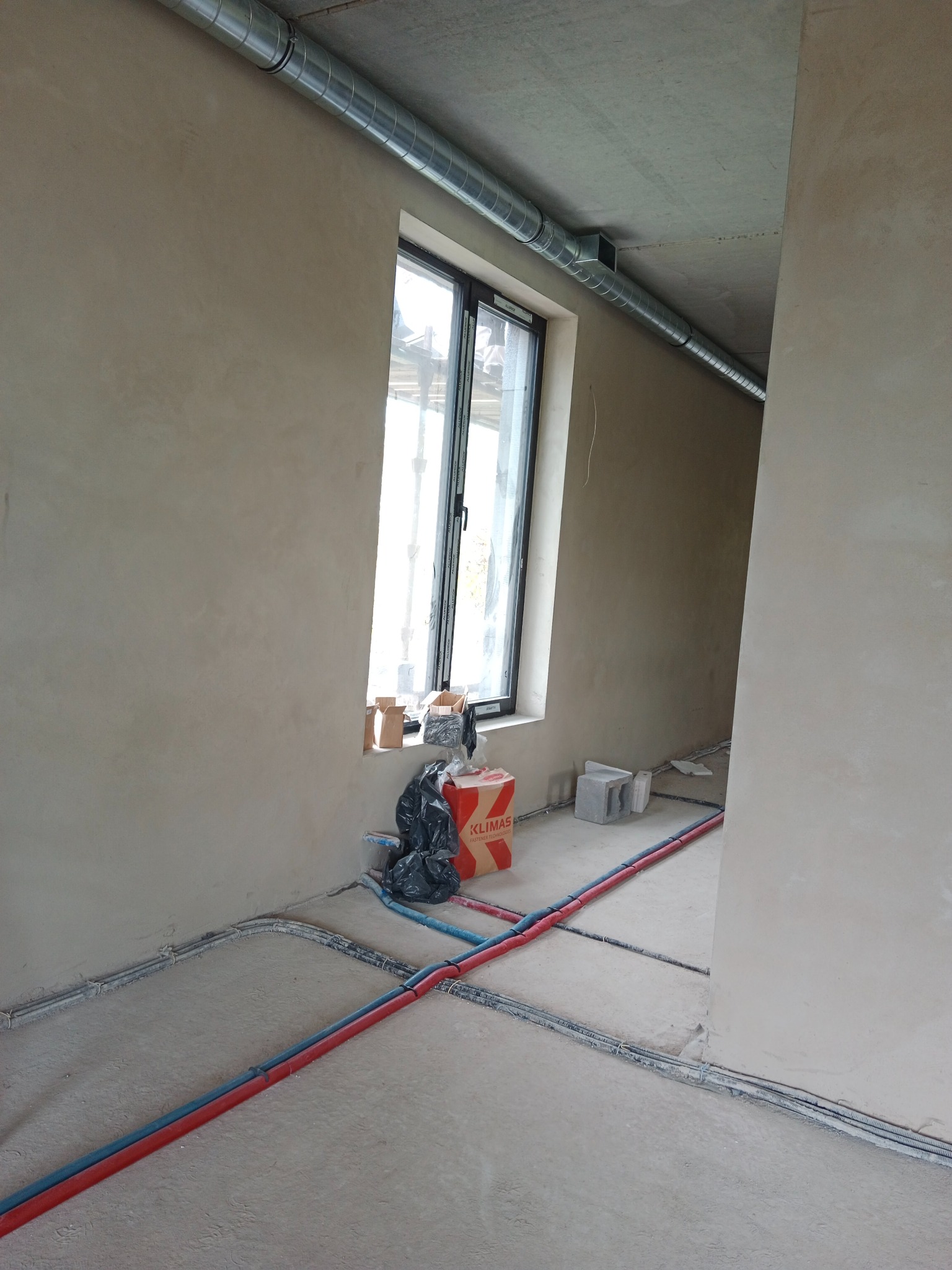 Surowy stan wnętrza z szarymi ścianami, widoczne rury instalacyjne w kolorze czerwonym i niebieskim na podłodze, oraz metalowa rura wentylacyjna pod sufitem, okno z widokiem na zewnątrz, kartonowe...